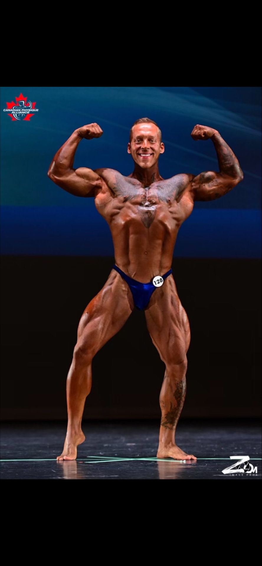 Colton Britzius hitting a front double biceps pose on the Canadian Physique Alliance stage, contestant number 128